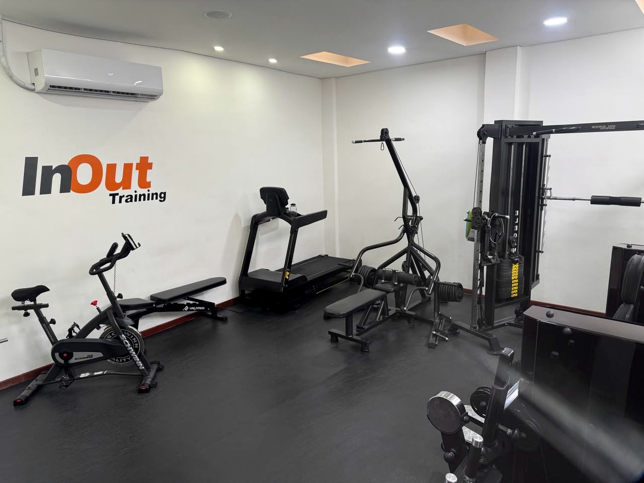 Visão ampla do salão do estúdio InOut Training: bicicleta, banco regulável, esteira e estação de musculação, com a marca aplicada na parede e ar-condicionado ao fundo.